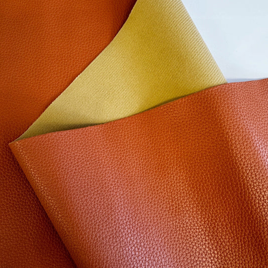 Close-up of an orange pebble-grained faux leather fabric showing a textured surface and a light yellow, soft backing
