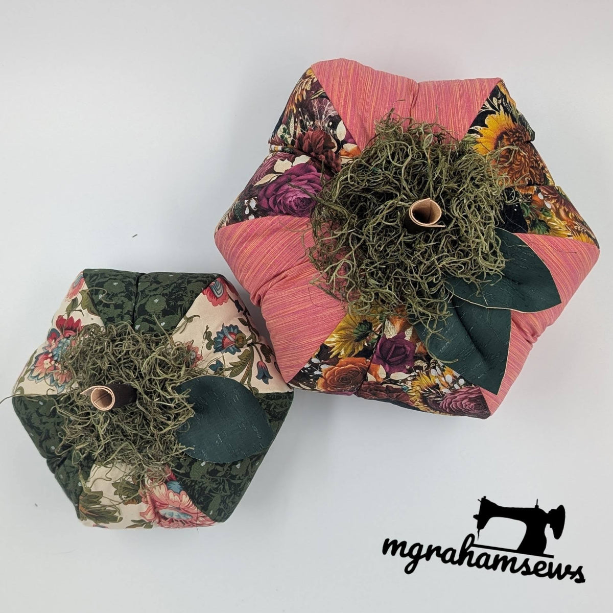 Two handmade patchwork fabric pumpkins featuring green, beige floral, rose, and autumn floral fabrics with moss-wrapped stems and green leaves