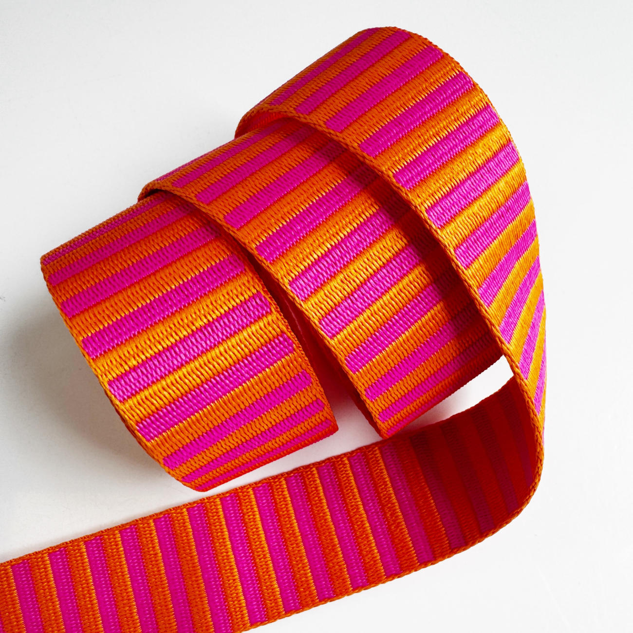 A 1.5-inch wide jacquard woven webbing strap showcasing a vibrant hot pink and orange horizontal stripe pattern, loosely coiled against a white background.