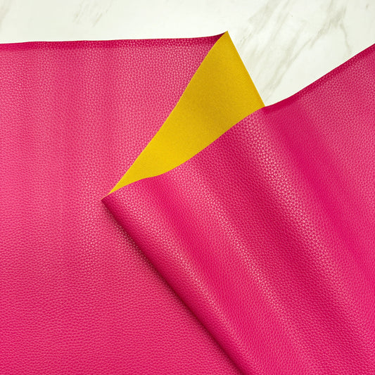 A sheet of vibrant, pink faux leather vinyl fabric with a finely pebbled texture. The top edge is folded down to reveal a stark, contrasting bright yellow backing material underneath. The fabric is casually draped, showcasing its flexible and supple quality against a light, grey-veined marble surface background.