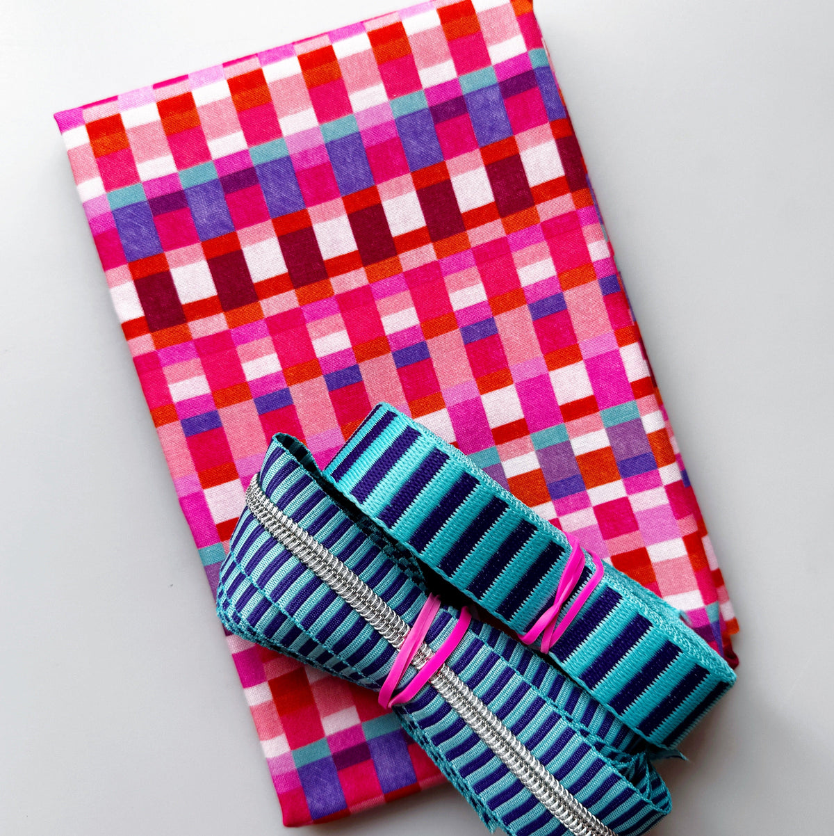 Pink gingham fabric with turquoise striped webbing and silver zipper for sewing and bag making.