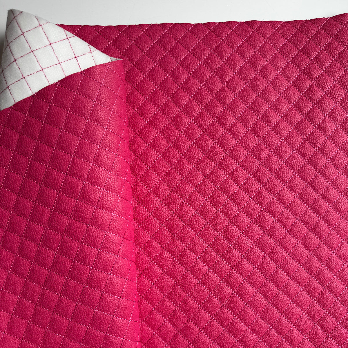 A close-up of a bright pink quilted fabric or cushion cover with a diamond stitched pattern, shown with one corner folded back to reveal a white reverse side with a red grid pattern. A hand is visible at the bottom left holding the fabric.