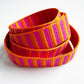 A coiled woven strap featuring alternating vertical stripes of bright orange, magenta pink, and red, photographed on a clean white background.