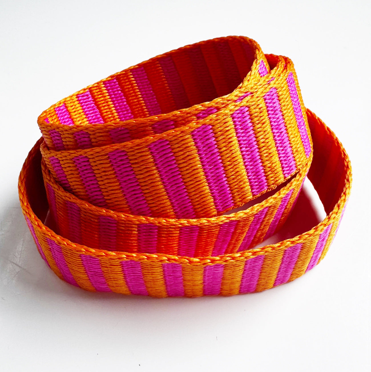 A coiled woven strap featuring alternating vertical stripes of bright orange, magenta pink, and red, photographed on a clean white background.