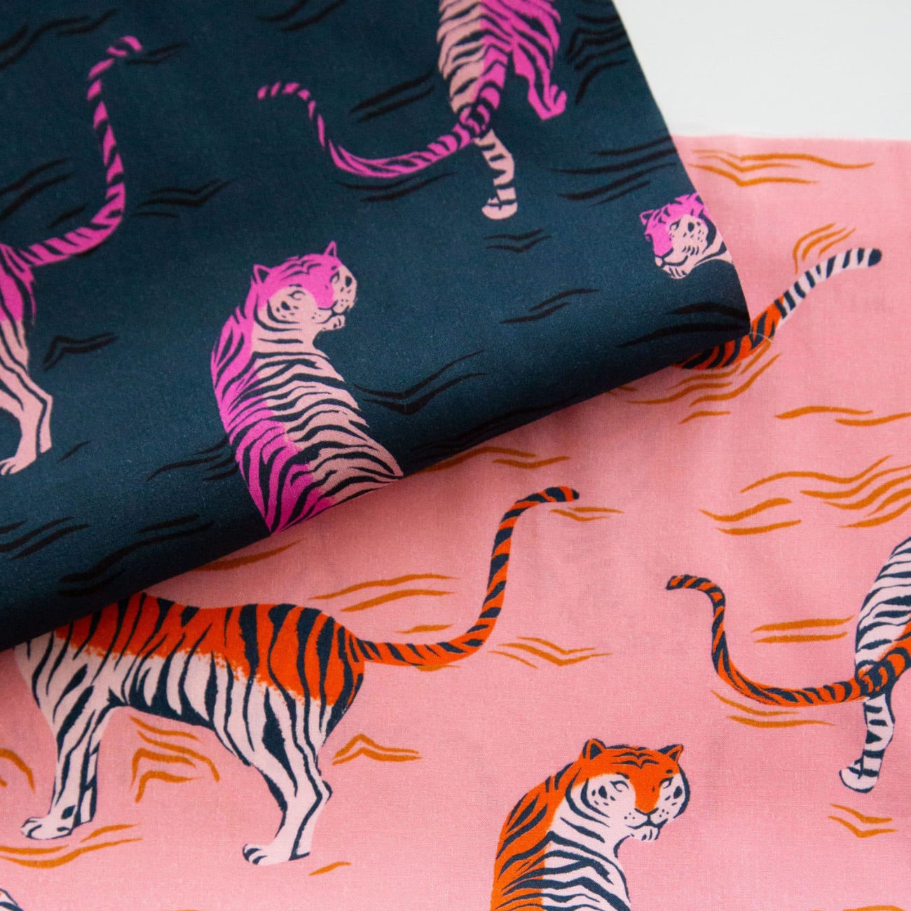 Two folded pieces of Ruby Star Society's Poolside Too Rayon fabric featuring a tiger print pattern. The top fabric is dark teal with pink and lavender tigers, and the bottom fabric is a sorbet pink with orange and white tigers swimming in water.