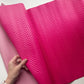 A close-up of a bright pink quilted fabric or mat being held open by a hand, showing a raised chevron pattern. A second pink item is partially visible underneath on a white surface.
