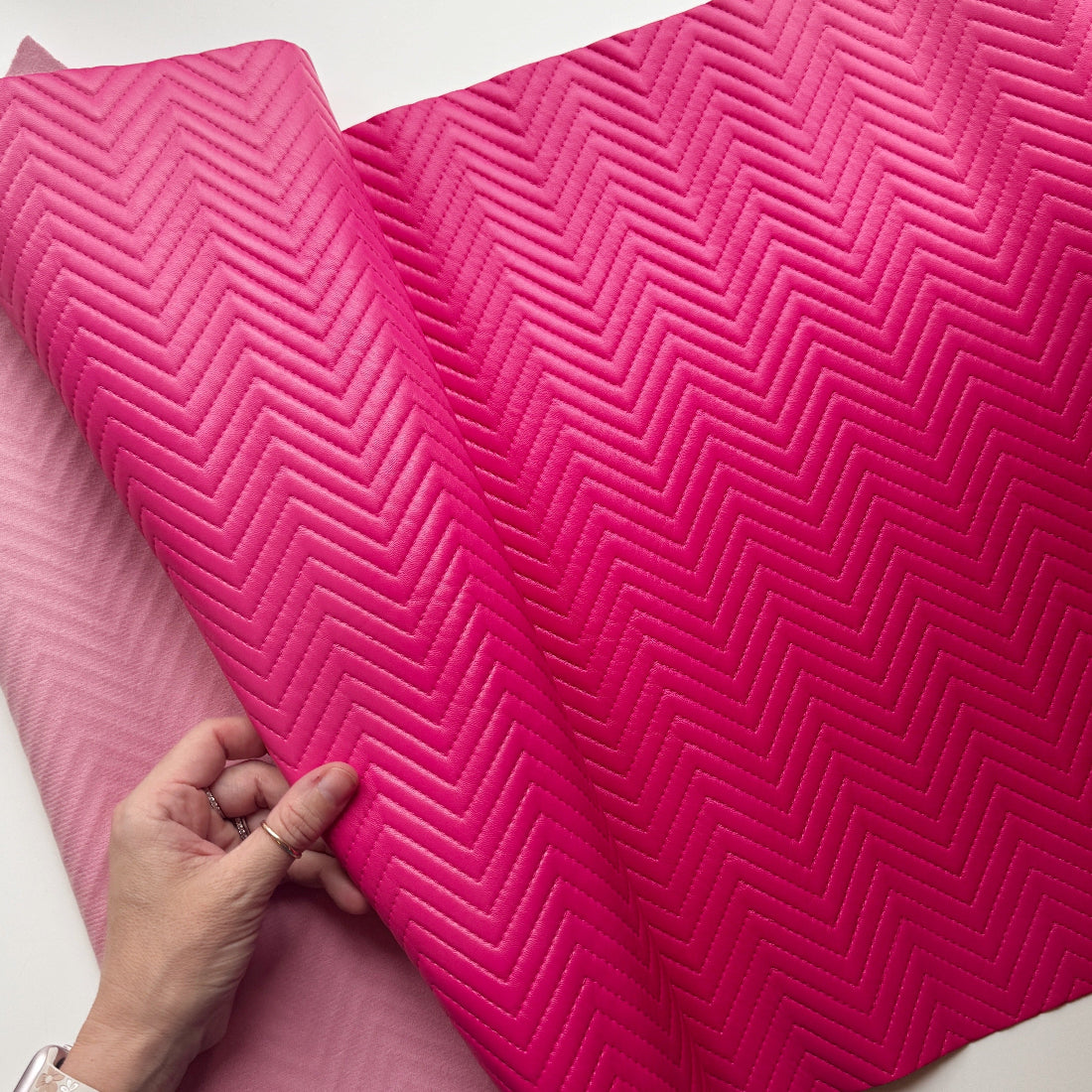 A close-up of a bright pink quilted fabric or mat being held open by a hand, showing a raised chevron pattern. A second pink item is partially visible underneath on a white surface.