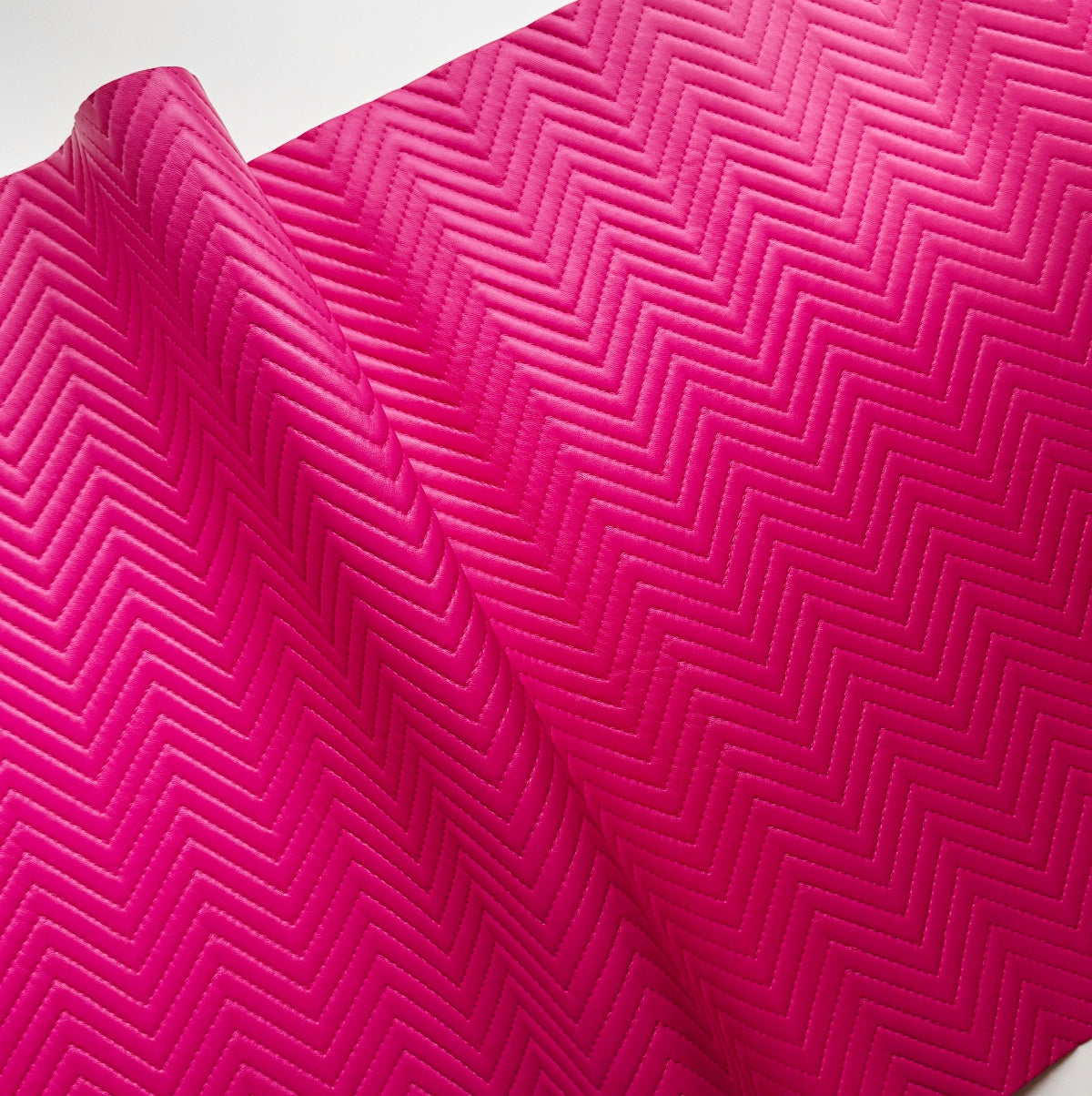 A close-up of a bright pink textured surface with a repeating chevron or zigzag embossed pattern, shown with a folded corner revealing a white background.