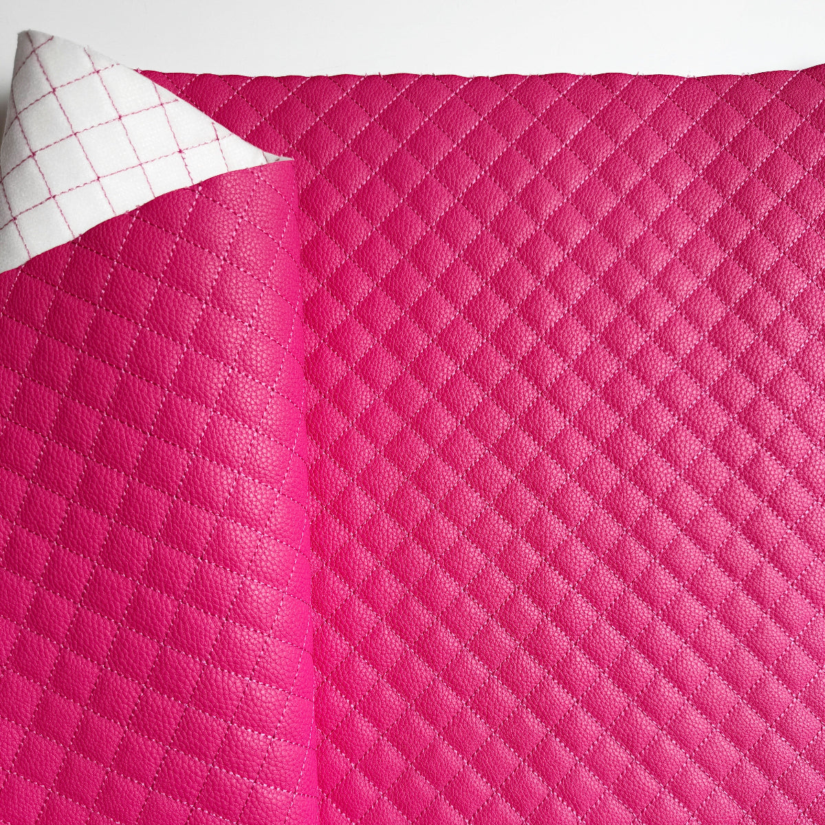 A close-up of a bright pink quilted fabric or leather-like material with a diamond stitched pattern, folded over to show a white backing with a pink grid pattern.