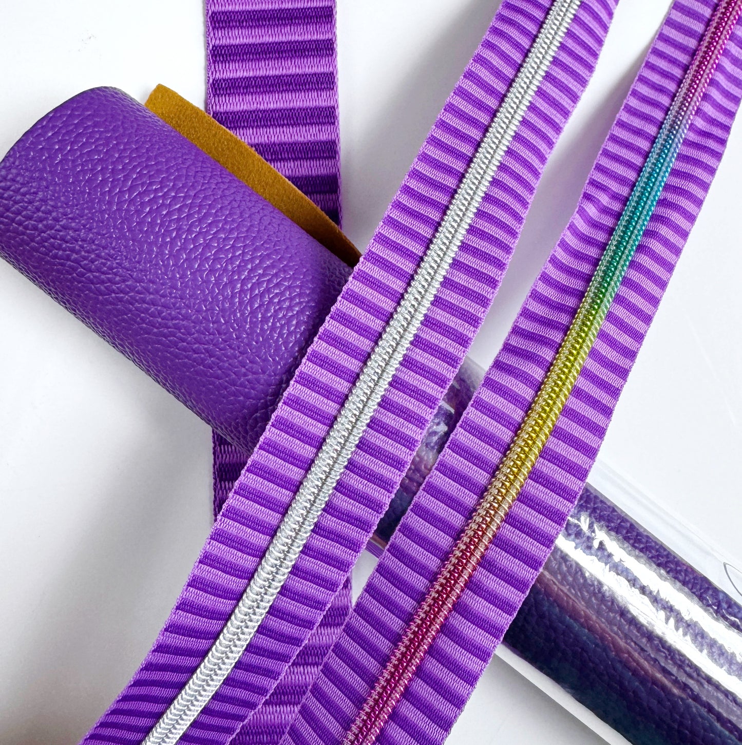 A purple themed bag making flat lay featuring a rolled piece of textured purple faux leather, a roll of translucent purple vinyl, a striped jacquard webbing strap, a matching zipper tape with silver coil teeth, and a second zipper tape with rainbow metallic teeth.