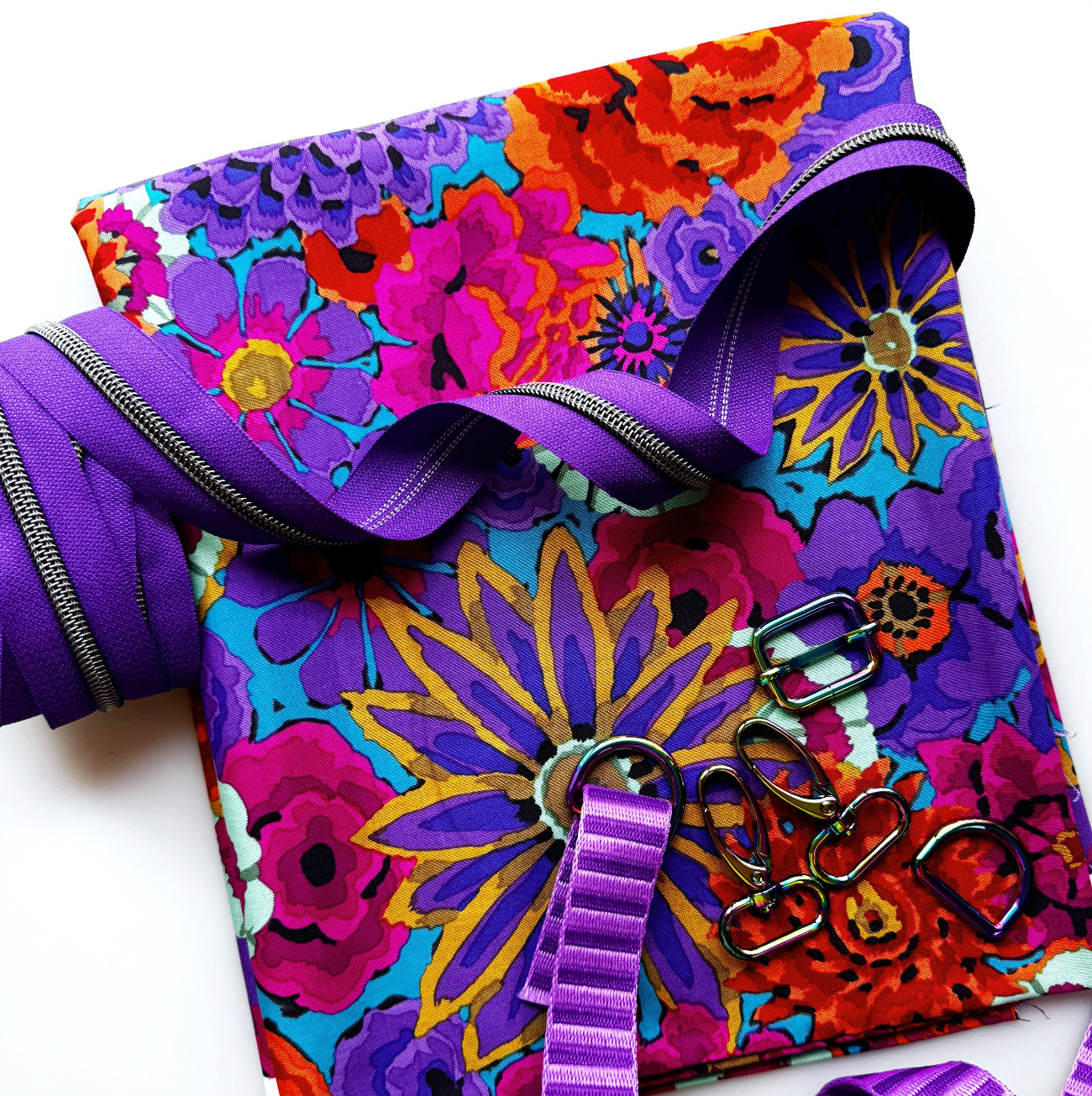 Vibrant purple floral sewing project featuring colorful fabric, purple striped jacquard webbing,  gunmetal zipper tape, and rainbow metal hardware.