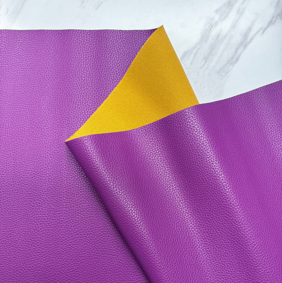 A close-up shot of a textured, pebbled faux leather vinyl fabric sheet. The main side is a vibrant orchid purple, and a corner is folded over to reveal a contrasting mustard-yellow backing material. The fabric is set against a white marble-patterned background.