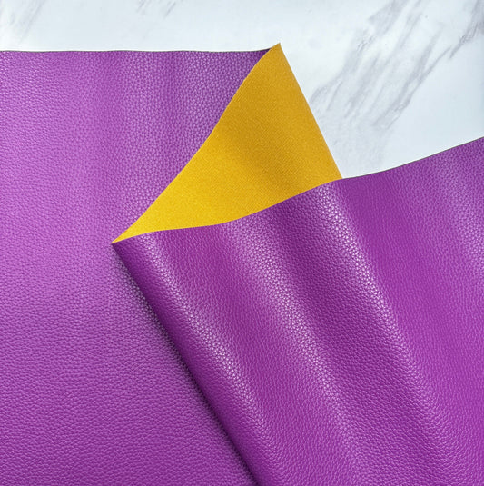 A close-up shot of a textured, pebbled faux leather vinyl fabric sheet. The main side is a vibrant orchid purple, and a corner is folded over to reveal a contrasting mustard-yellow backing material. The fabric is set against a white marble-patterned background.