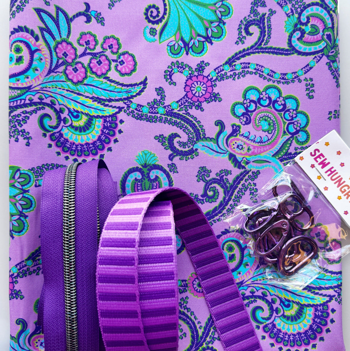 Purple paisley fabric with coordinating purple webbing, zipper tape, and metal bag hardware for a sewing project.