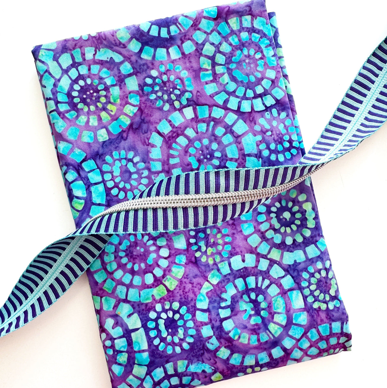 Folded purple and teal batik fabric with a circular mosaic pattern, paired with a coordinating blue and purple striped zipper tape with silver teeth.