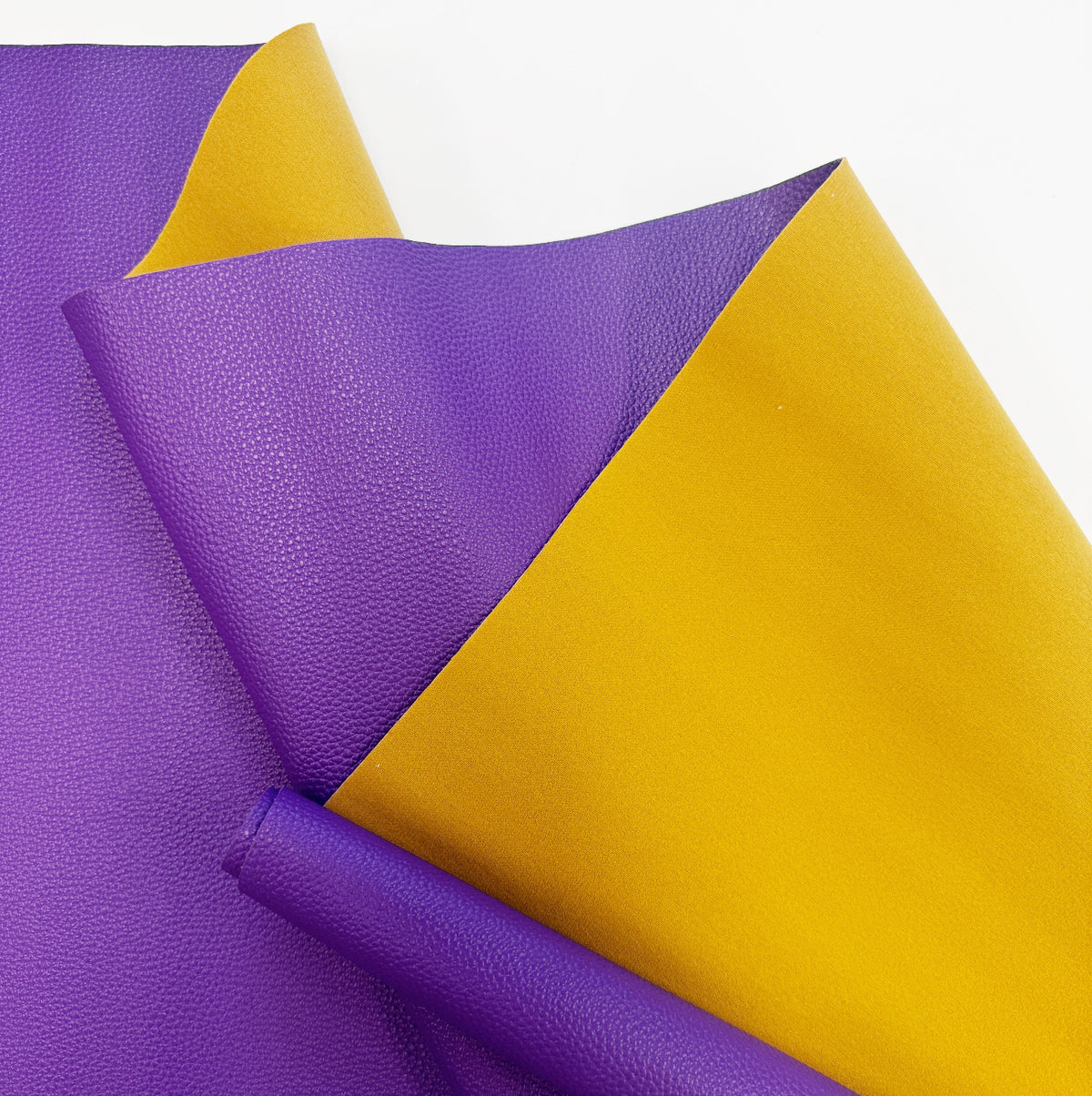 A close-up shot of dual-sided faux leather fabric featuring a textured, vibrant purple side and a smooth, golden-yellow reverse side, draped in loose folds against a white background