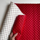A close-up of a red quilted faux-leather or leather-like mat being held by a hand, with the underside showing a white grid pattern. The product is photographed against a plain light background.