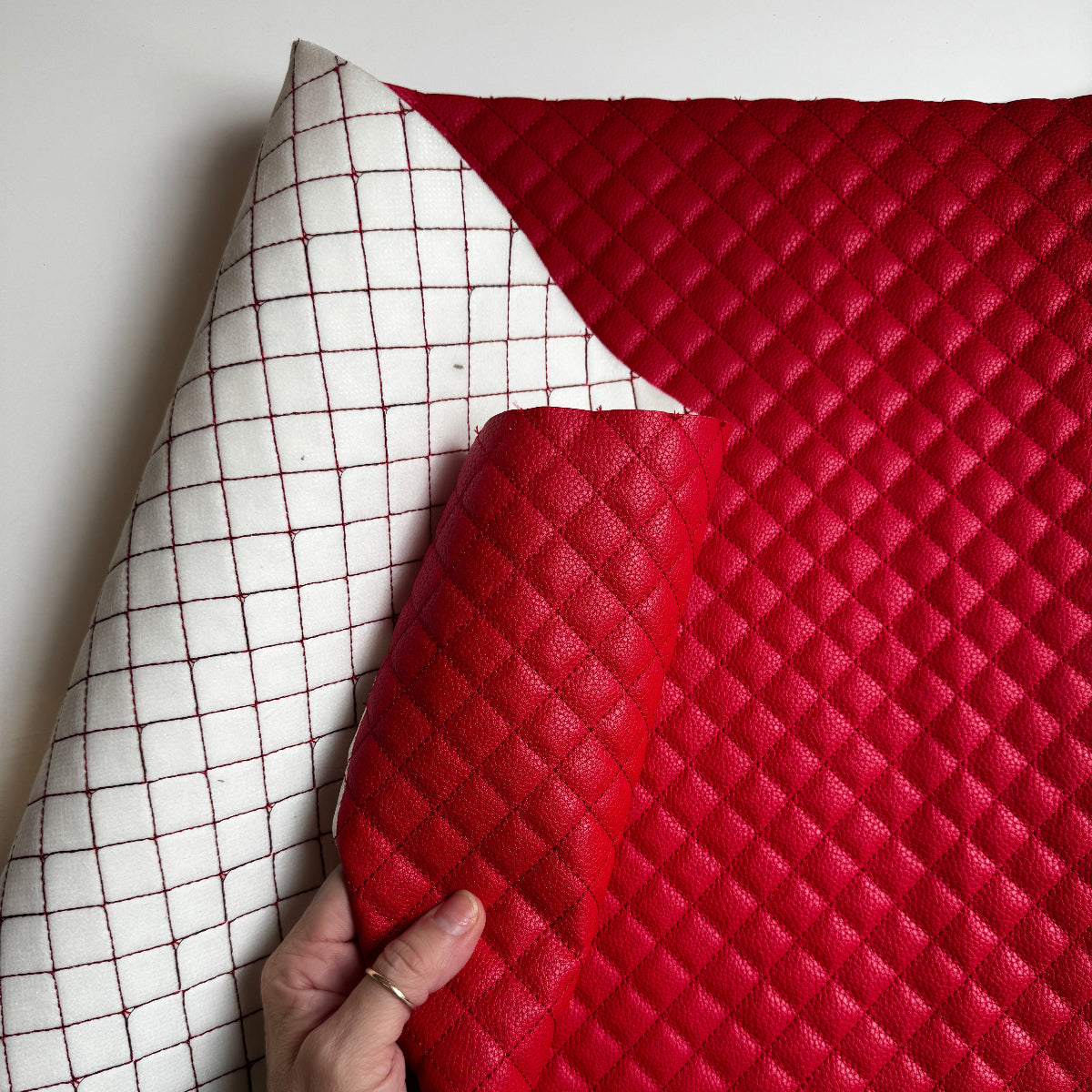 A close-up of a red quilted faux-leather or leather-like mat being held by a hand, with the underside showing a white grid pattern. The product is photographed against a plain light background.