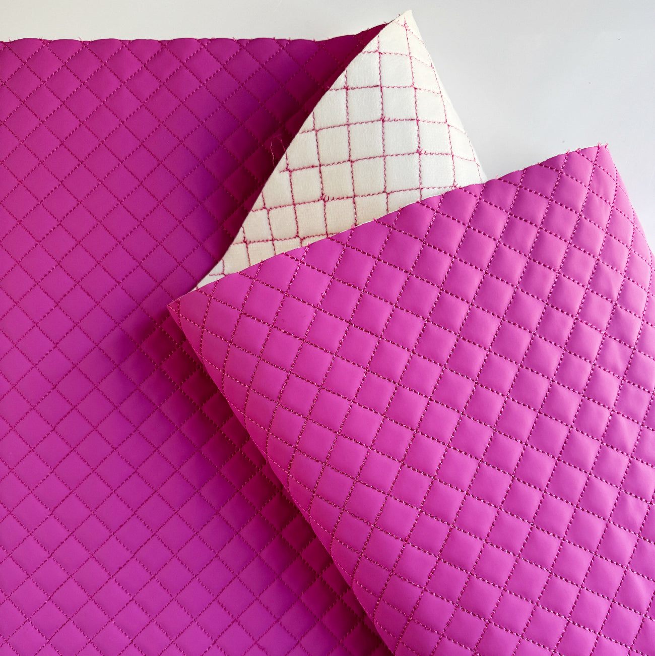 A folded quilted fabric item in bright pink with a diamond stitched pattern, shown against a light gray background. The underside reveals a white backing with a pink grid pattern.