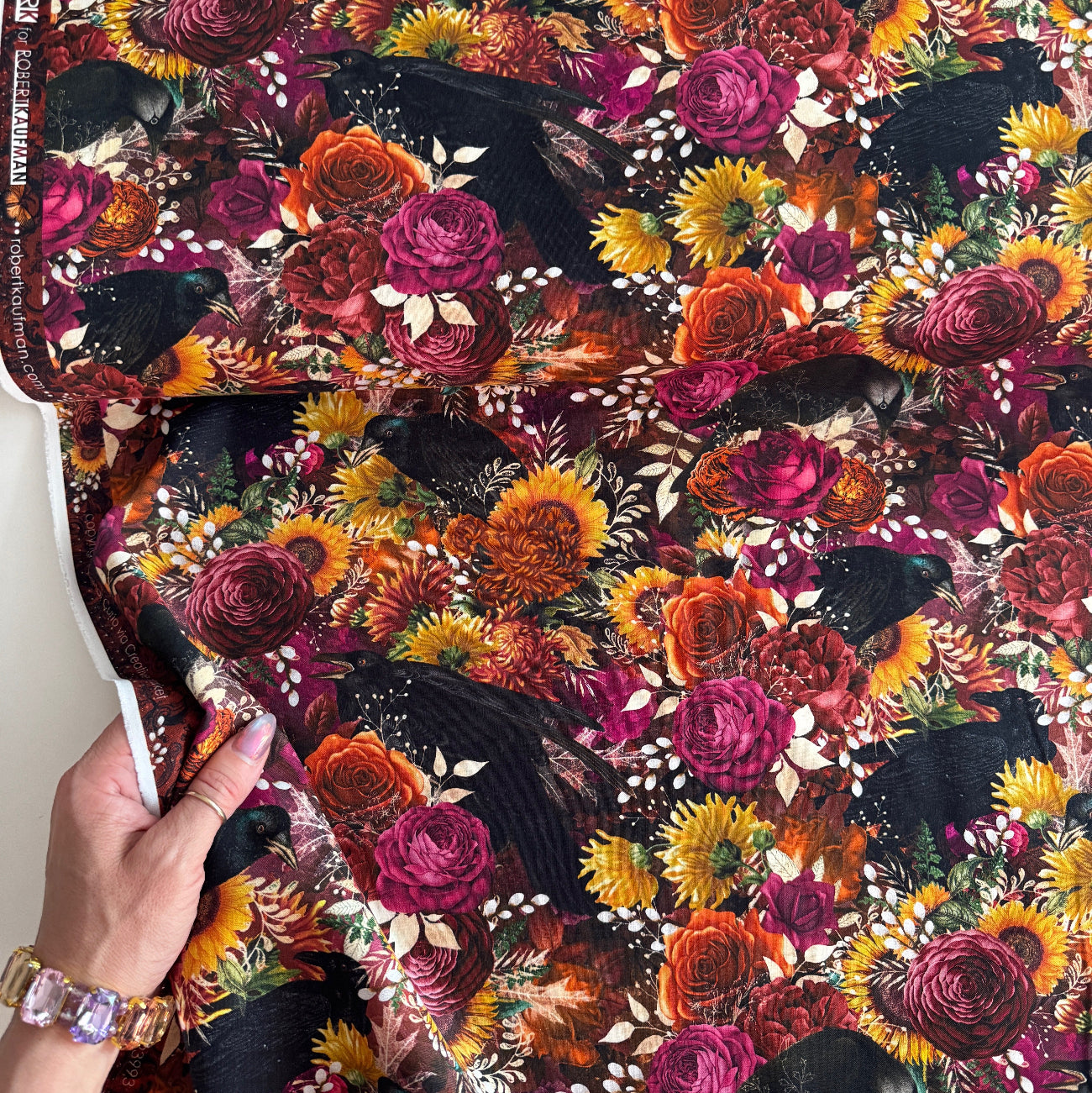 Hand holding a fabric "Ravenwood" fabric  showing the "Eerie" pattern dark, maximalist floral design with black crows, sunflowers, and vibrant roses.