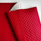 A close-up of a red quilted faux leather or leather fabric sheet with a diamond stitched pattern, folded over to show its texture and a white backing with a red grid pattern.