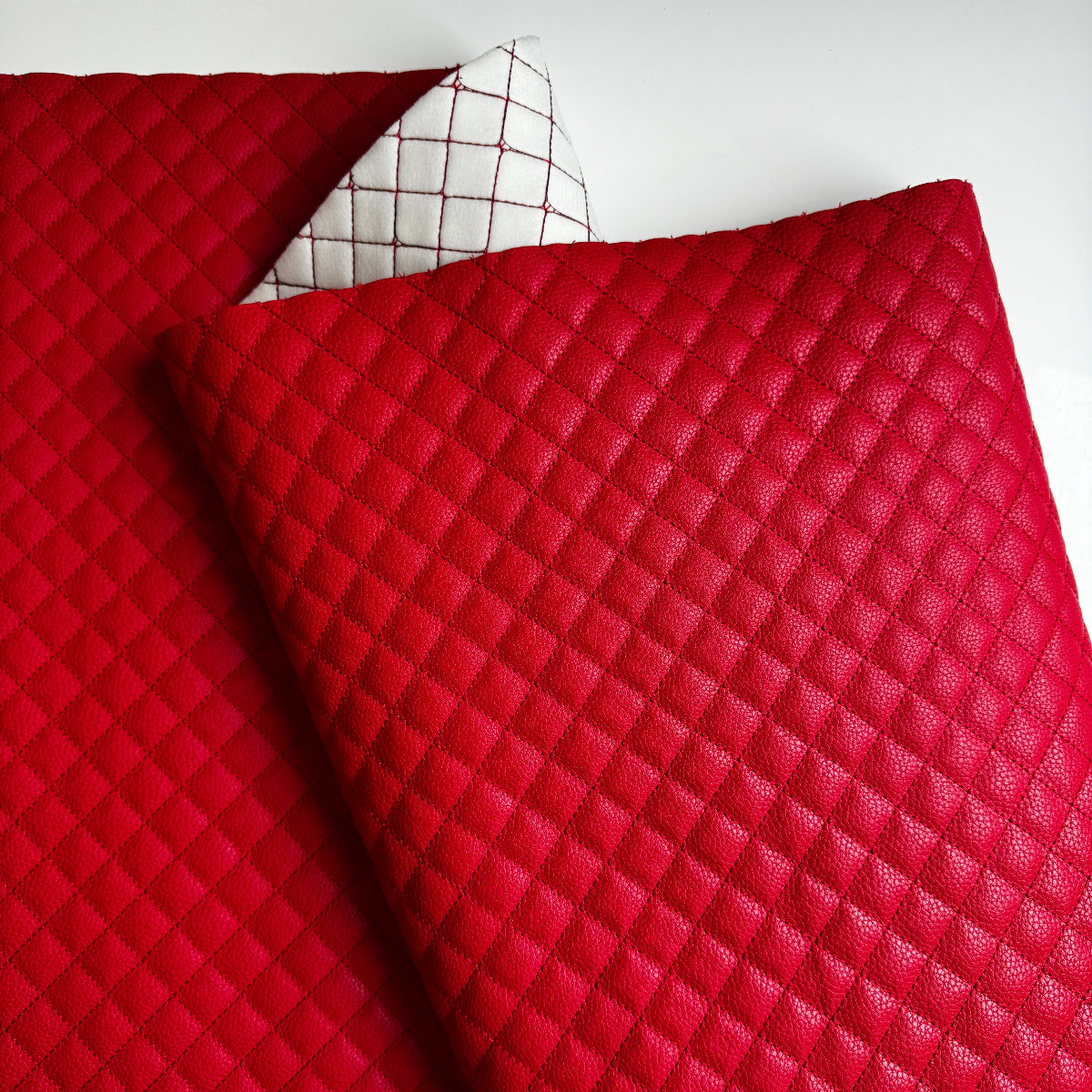 A close-up of a red quilted faux leather or leather fabric sheet with a diamond stitched pattern, folded over to show its texture and a white backing with a red grid pattern.