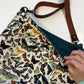 A close-up of a fabric shoulder bag with a dense vintage-style butterfly and moth print on an off-white background, featuring a dark teal interior lining and a brown leather strap. A hand is visible holding the bag's edge.