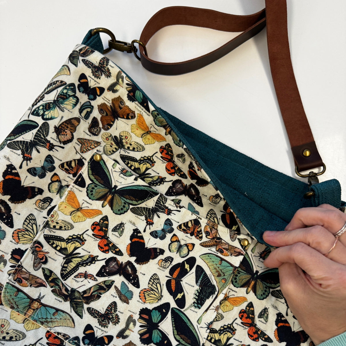 A close-up of a fabric shoulder bag with a dense vintage-style butterfly and moth print on an off-white background, featuring a dark teal interior lining and a brown leather strap. A hand is visible holding the bag's edge.
