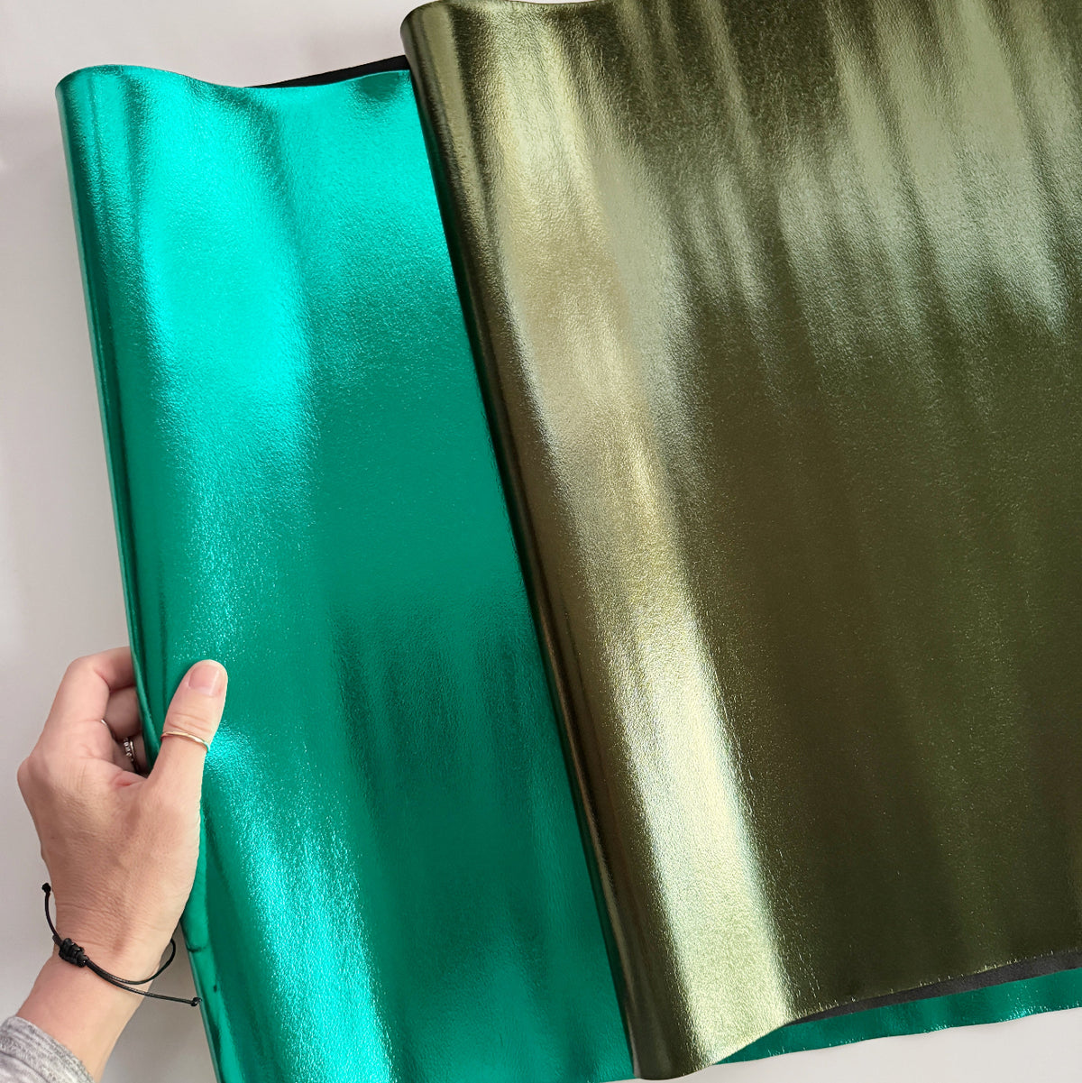 Iridescent metallic PU faux leather fabric in glossy teal and olive green with a reflective, color-shifting finish. Eye-catching vinyl material for handmade handbags, purses, wallets, accessories, and DIY bag-making projects.
