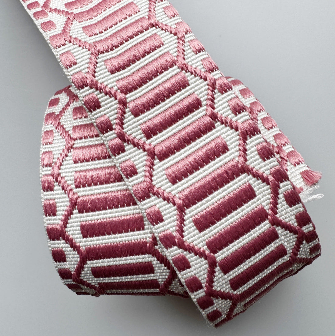 A close-up of a woven elastic strap with a geometric pattern in dusty pink and white, shown on a neutral light gray background.