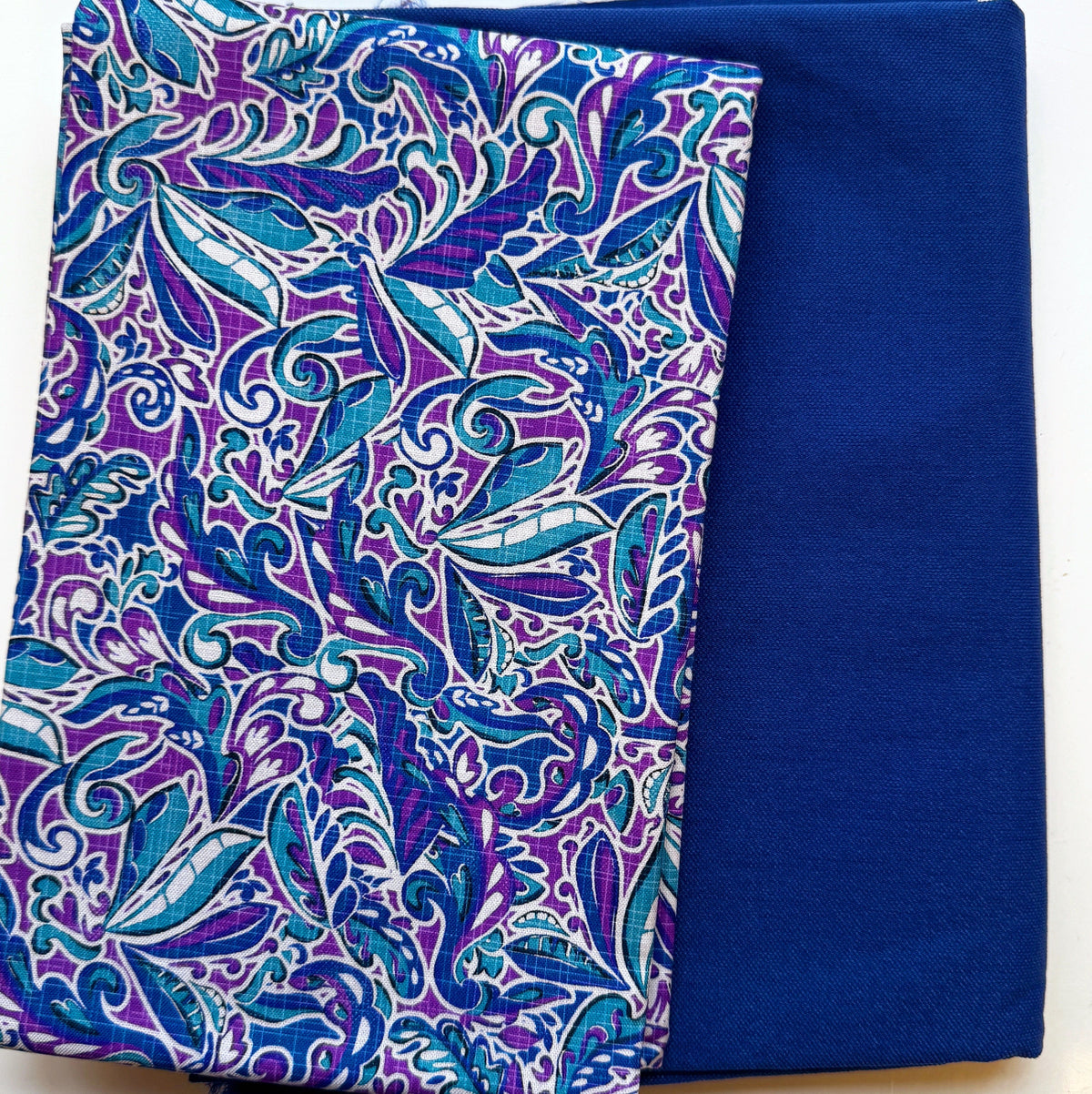 Folded quilting fabrics featuring a royal blue, purple, teal, and aqua leaf print paired with coordinating solid royal blue fabric