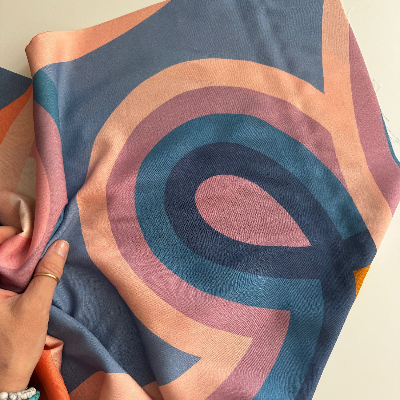 A person’s hand holds a draped piece of Ruby Star Society rayon fabric, featuring a modern, abstract pattern of bold, interlocking curved lines in shades of blue, peach, purple, and deep navy.