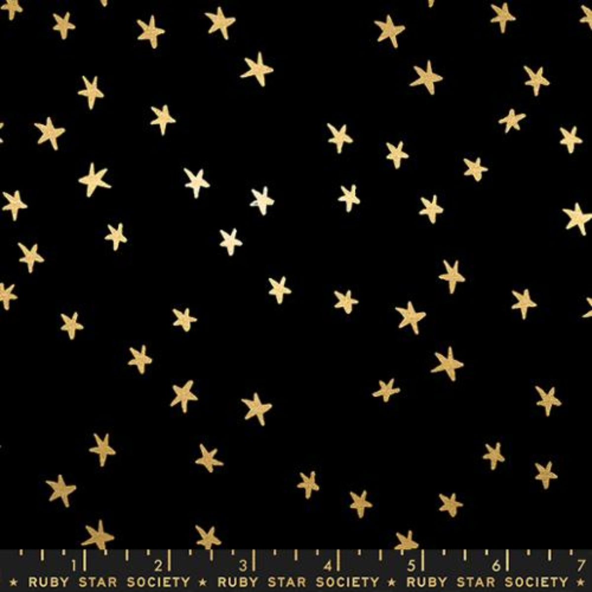 A flat lay photograph of a black fabric pattern featuring scattered, hand-drawn metallic gold stars of varying sizes. A quilting ruler at the bottom edge marks measurements in inches from 1 to 7, displaying the text "RUBY STAR SOCIETY" printed repeatedly in gold.