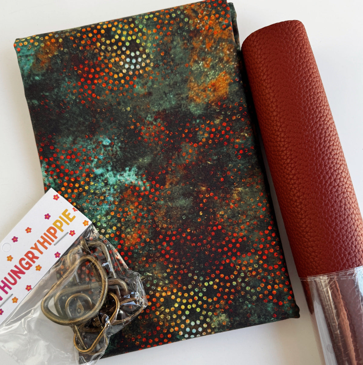 Crafting kit featuring a dark, patinated fabric with a metallic dot pattern Moda Tranquil Patina, a roll of rust-brown faux leather, and hardware labeled by Hungry Hippie
