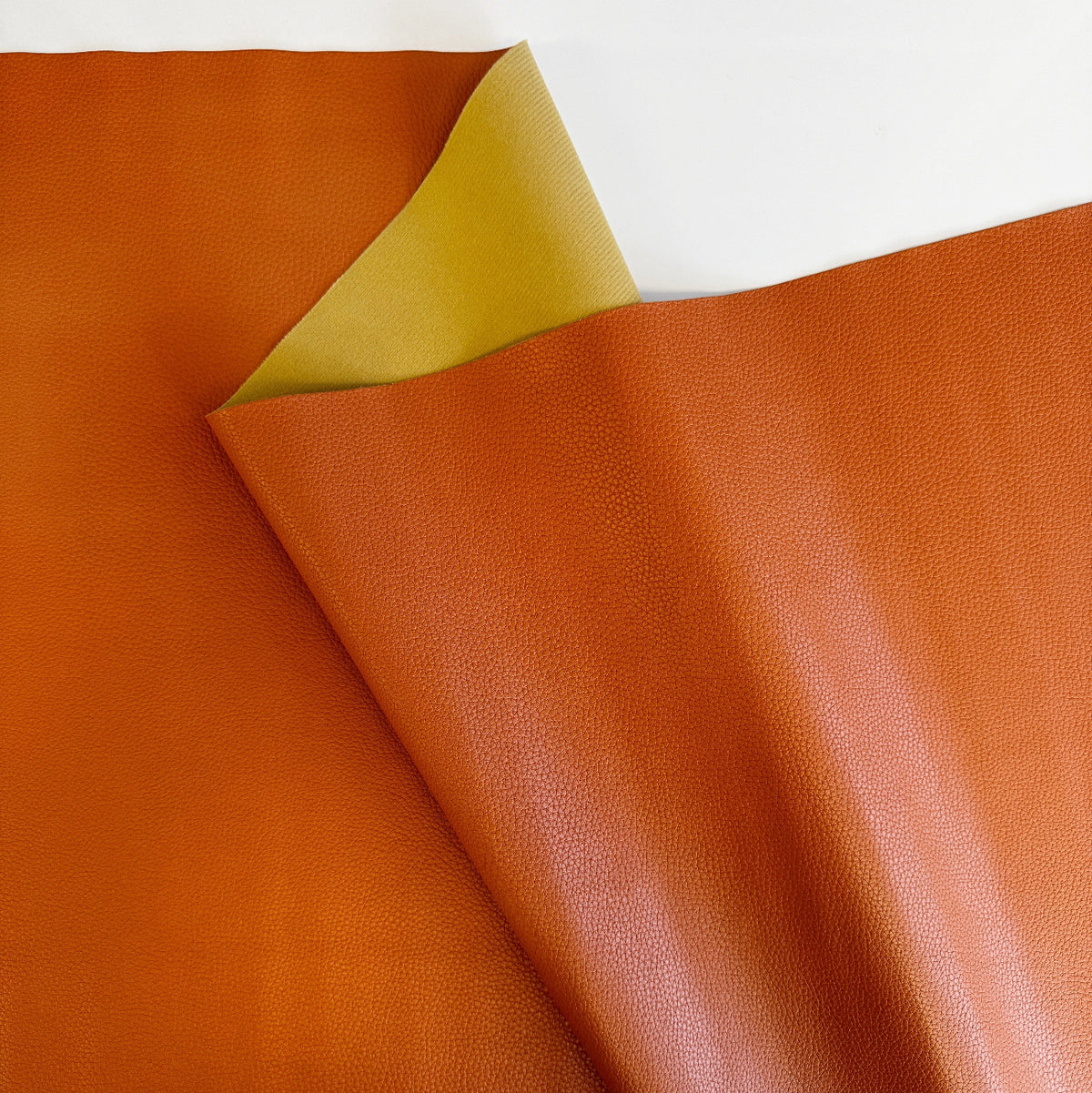 Rust orange textured faux leather fabric roll closeup, yellow soft backing on a white background