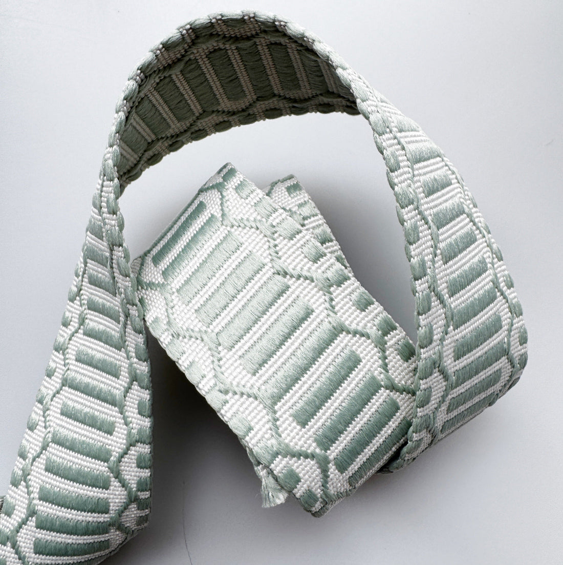 A light sage green and white woven fabric belt with a geometric pattern, folded and displayed on a light gray background.