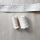 Two spools of Gutermann beige polyester sewing thread and a geometrically patterned cream-colored trim strip rest on top of a textured, champagne-colored faux leather surface.
