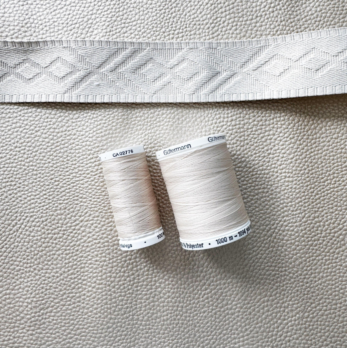 Two spools of Gutermann beige polyester sewing thread and a geometrically patterned cream-colored trim strip rest on top of a textured, champagne-colored faux leather surface.
