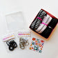 Sew Hungry Hippie sewing hardware and bag-making kit arranged on a white background, featuring black D-rings, silver metal D-rings and swivel clasps in clear retail packages, rolled zipper tape or straps in black, black-and-white stripes, and pink heart prints, plus a clear plastic storage container and a retro “Stay Groovy with Sewing” card. Sewing supplies for handmade bags, purses, and DIY accessories.