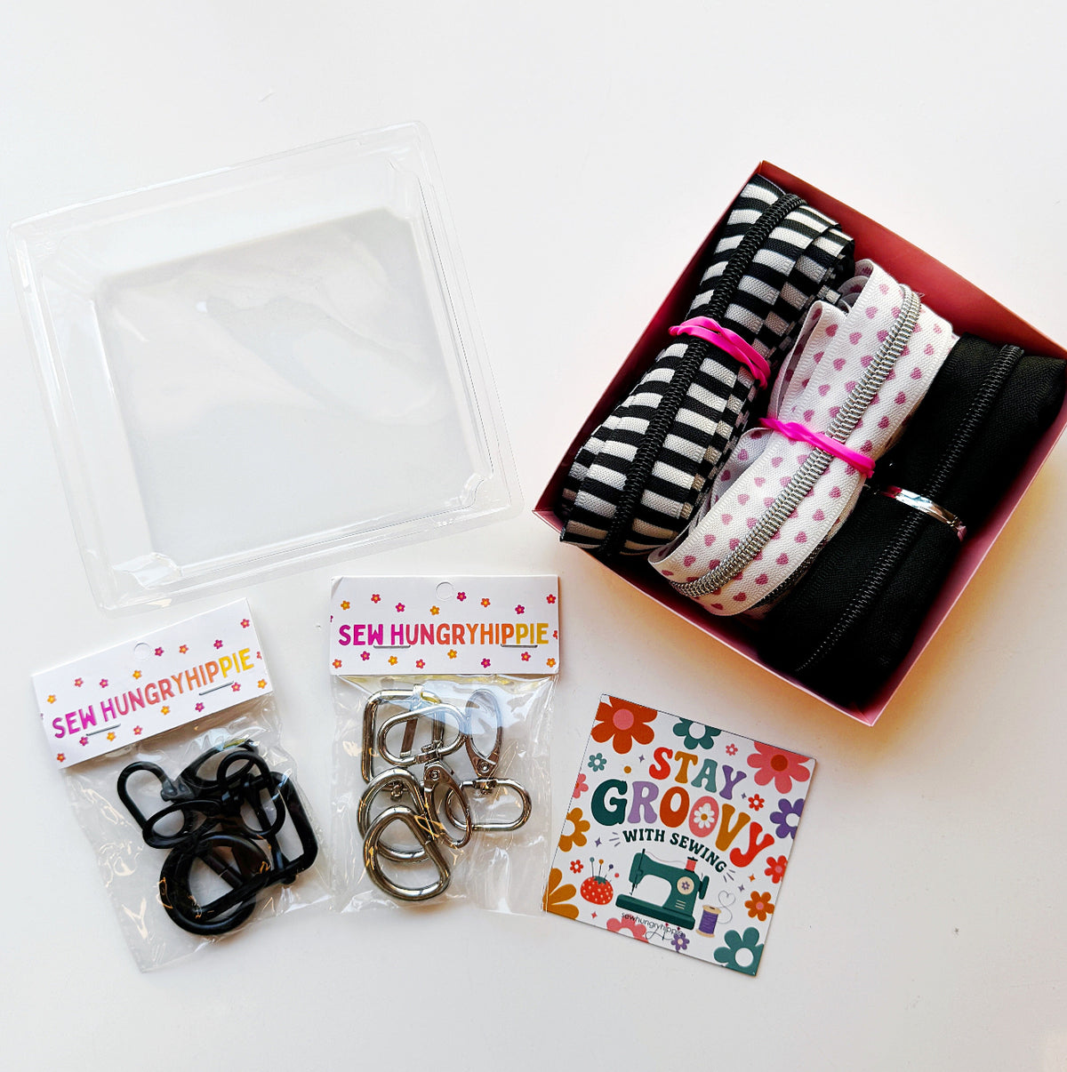 Sew Hungry Hippie sewing hardware and bag-making kit arranged on a white background, featuring black D-rings, silver metal D-rings and swivel clasps in clear retail packages, rolled zipper tape or straps in black, black-and-white stripes, and pink heart prints, plus a clear plastic storage container and a retro “Stay Groovy with Sewing” card. Sewing supplies for handmade bags, purses, and DIY accessories.