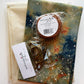 Sew Hungry Hippie DIY bag-making kit featuring cream cotton fabric, colorful galaxy-print fabric with blue, green, orange, and white splatters, a light brown 1.25-inch sewable leather bag strap, and antique brass slider buckles and swivel snap hooks. Complete bag-making supplies for handmade handbags, purses, and sewing projects.