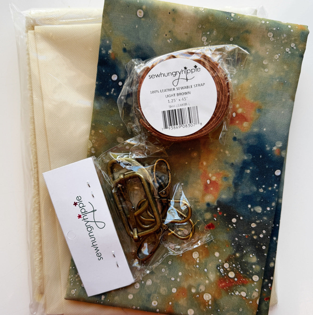 Sew Hungry Hippie DIY bag-making kit featuring cream cotton fabric, colorful galaxy-print fabric with blue, green, orange, and white splatters, a light brown 1.25-inch sewable leather bag strap, and antique brass slider buckles and swivel snap hooks. Complete bag-making supplies for handmade handbags, purses, and sewing projects.