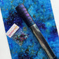 A roll of iridescent purple and black crocodile-embossed faux leather fabric lies diagonally across a vibrant blue and purple galaxy-print fabric background. Next to the rolled fabric, a small plastic baggie contains iridescent rainbow-colored metal D-rings, featuring a "Sew Hungry Hippie" branded paper label wrapper at the top.