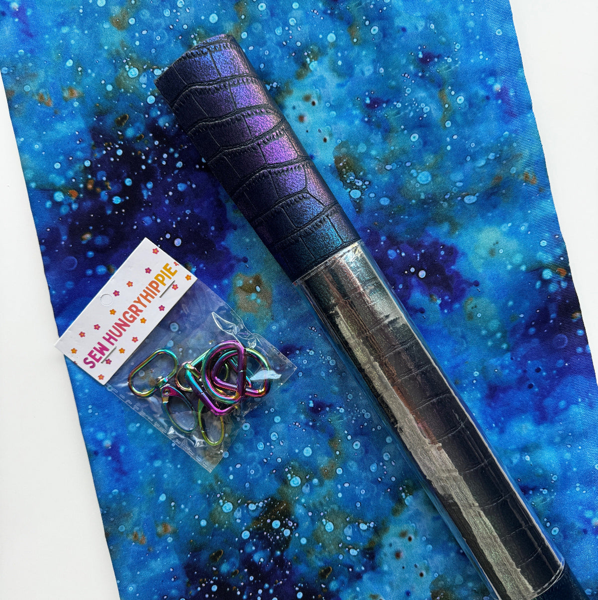 A roll of iridescent purple and black crocodile-embossed faux leather fabric lies diagonally across a vibrant blue and purple galaxy-print fabric background. Next to the rolled fabric, a small plastic baggie contains iridescent rainbow-colored metal D-rings, featuring a "Sew Hungry Hippie" branded paper label wrapper at the top.