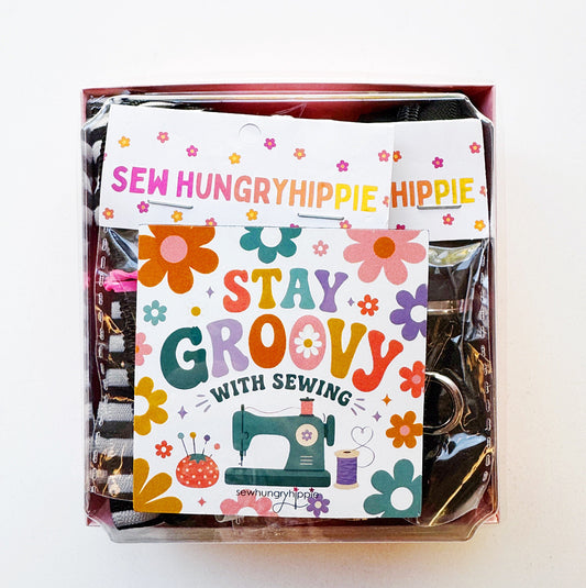 Sew Hungry Hippie bag-making hardware kit flat lay featuring snack pack zippers, magnets, black D-rings, silver metal D-rings and swivel clasps, and rolled zipper tape or straps in black, black-and-white stripes, and pink heart prints. Includes a clear plastic storage container, pink box, and colorful retro “Stay Groovy with Sewing” card decorated with 1970s-style flowers and a vintage sewing machine. Sewing supplies for handmade handbags, zipper pouches, purses, and DIY accessories.