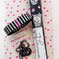 Top-down view of a bag-making supply bundle from "Sew Hungry Hippie" resting on white fabric with small pink stars. The kit includes a rolled piece of black textured vinyl decorated with colorful sugar skulls and flowers, a bundle of black-and-white vertically striped strap webbing, and a small plastic bag containing black metal bag hardware, including sliders and swivels. A white label with black script text reading "sewhungryhippie" is wrapped around the vinyl roll.