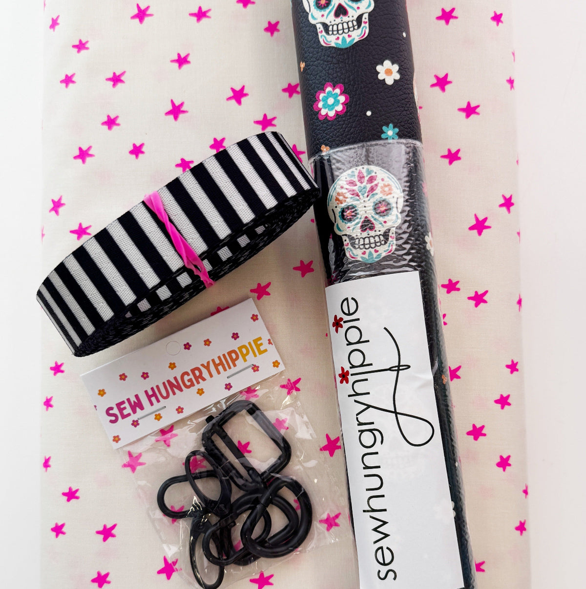 Top-down view of a bag-making supply bundle from "Sew Hungry Hippie" resting on white fabric with small pink stars. The kit includes a rolled piece of black textured vinyl decorated with colorful sugar skulls and flowers, a bundle of black-and-white vertically striped strap webbing, and a small plastic bag containing black metal bag hardware, including sliders and swivels. A white label with black script text reading "sewhungryhippie" is wrapped around the vinyl roll.