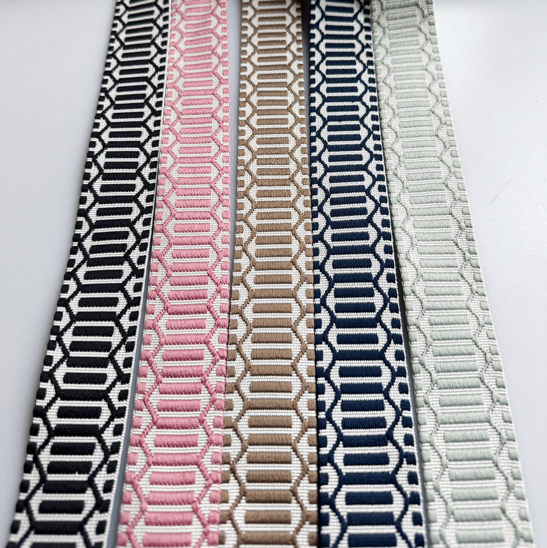 Five woven elastic straps with geometric patterns are arranged vertically side-by-side on a white background. The straps feature black and white, pink and white, tan and white, navy and white, and light gray and white patterns.