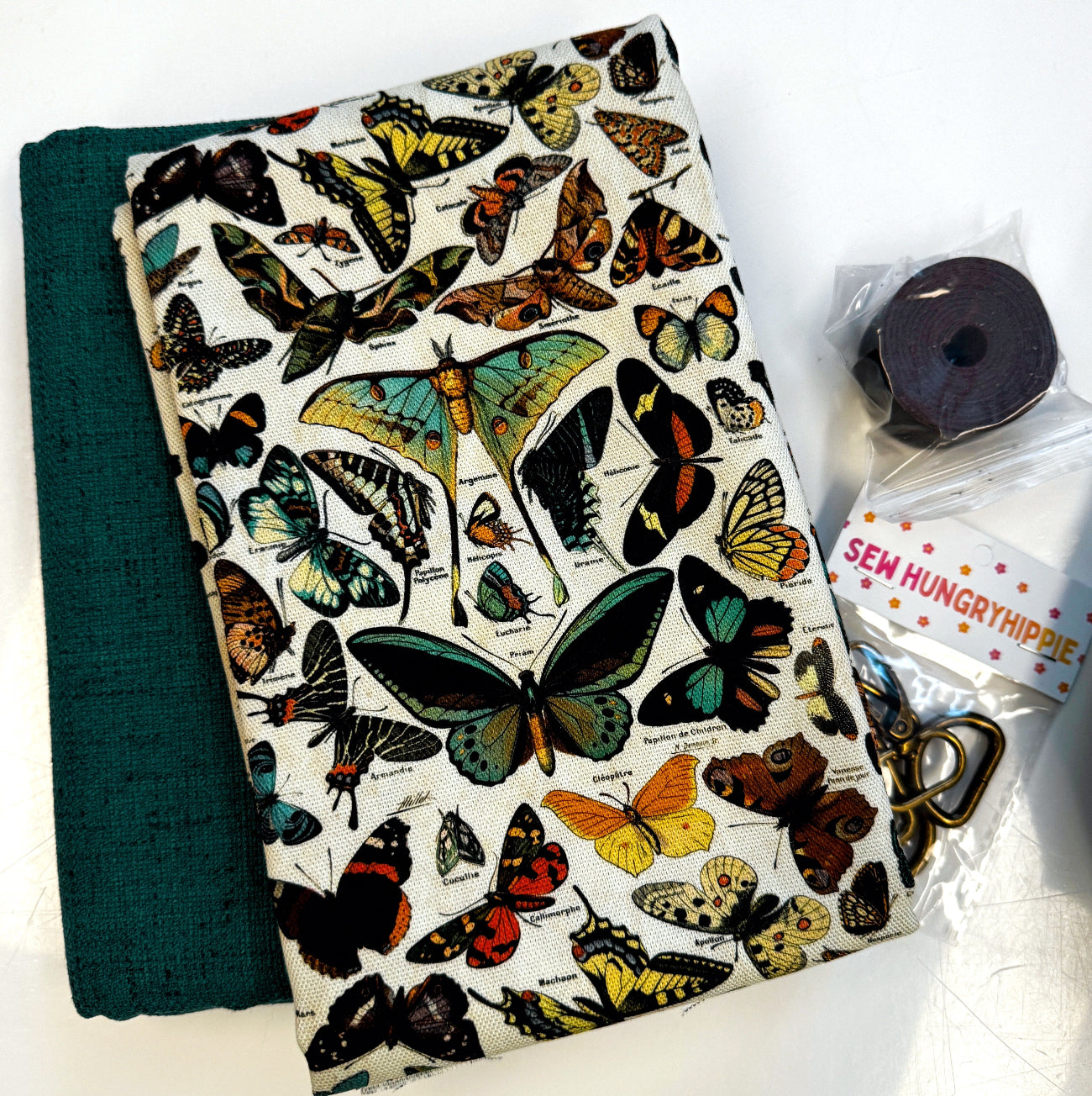 A fabric zippered pouch with a vintage-style butterfly print on a cream background, displayed next to a folded dark green fabric piece and a small packaged sewing thread spool with a tag reading 'SEW HUNGRIPHIDE'.