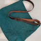 A teal woven fabric shoulder bag with a brown leather strap and brass buckle, laid flat on a white surface.
